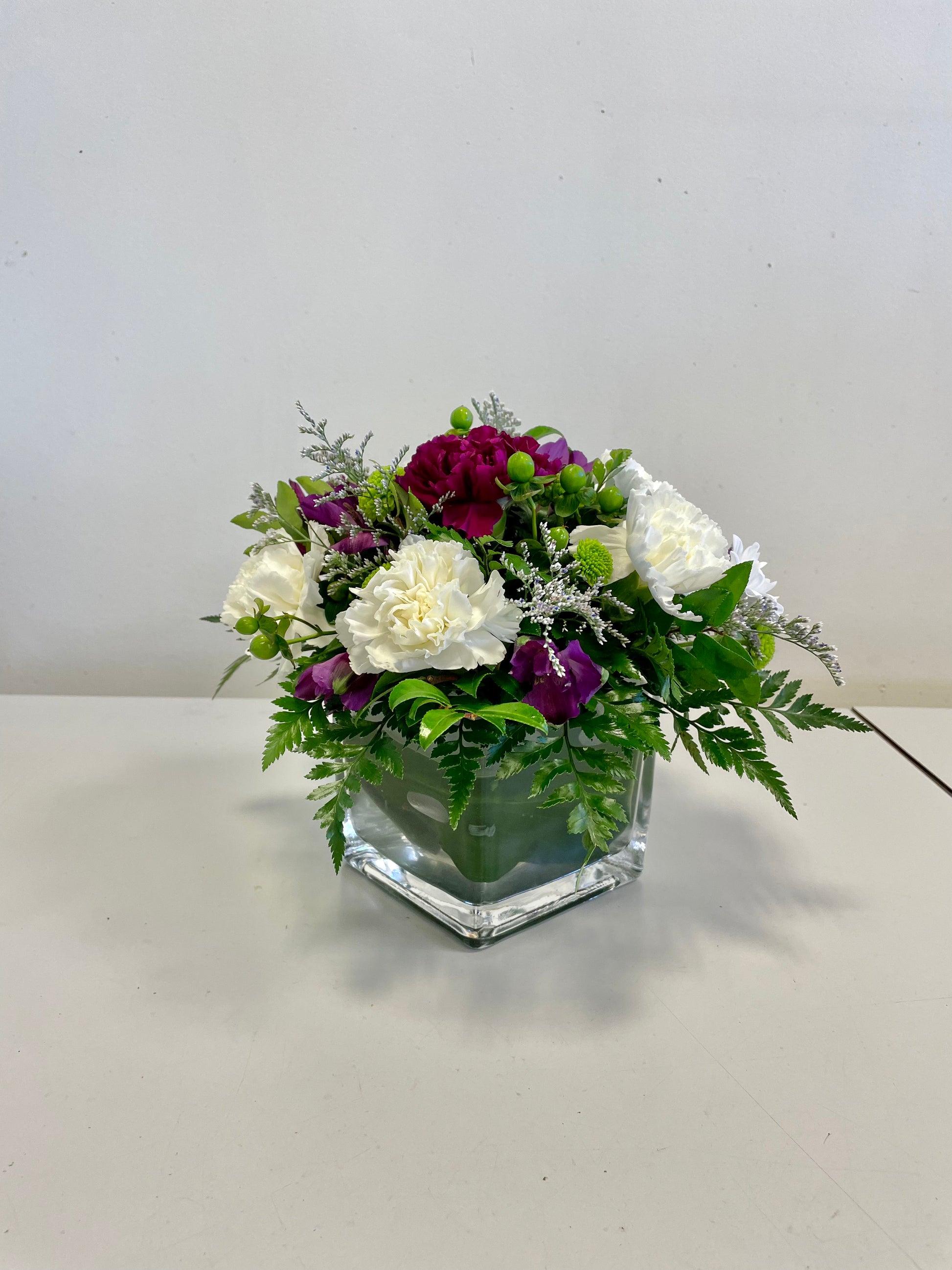 The Beloved Bouquet from Posies Flowers & Fashion is a floral arrangement in a clear square glass vase, featuring a mix of purple, white, and green flowers, including carnations and chrysanthemums, interspersed with green foliage and delicate white fillers. Due to seasonal availability, occasional substitutions may occur. The vase is placed on a white surface against a plain background.