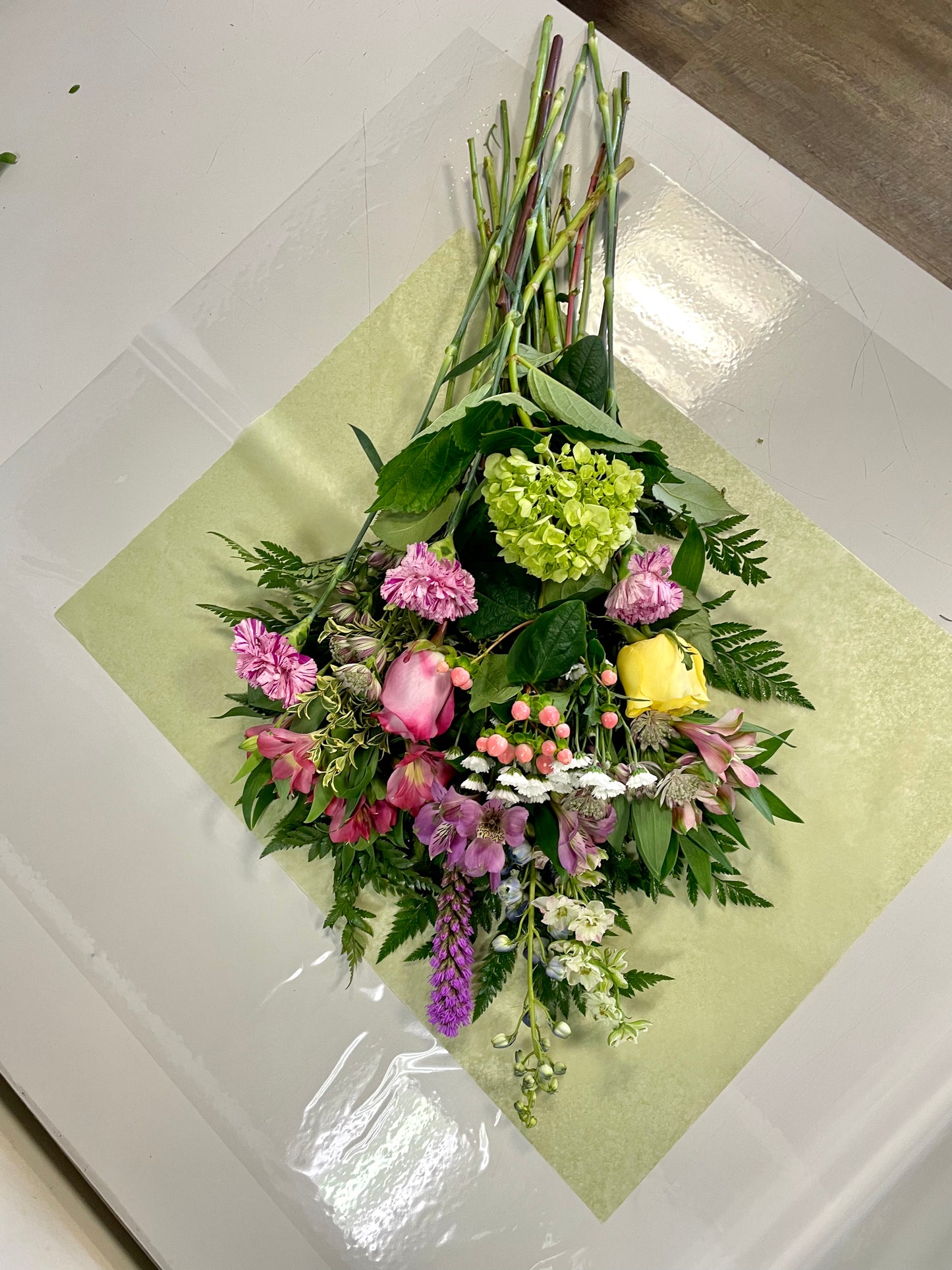 On a table lies a vibrant floral arrangement named "Acknowledge Worthy" by Posies Flowers & Fashion. This stunning bouquet features an exquisite variety of flowers including pink tulips, yellow roses, white daisies, purple liatris, and more, all complemented by lush green foliage. The flowers are beautifully displayed on light green paper, creating an image of pure elegance. Please note that the availability of flowers may vary.