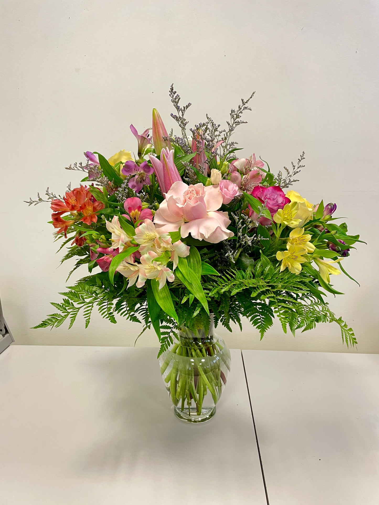 The Summer Serenade by Posies Flowers & Fashion features a clear glass vase filled with a vibrant bouquet of mixed flowers, including pink lilies, yellow and pink alstroemerias, and various green foliage. The arrangement is showcased against a plain white background on a white table. Please note that substitutions may be made based on flower availability.