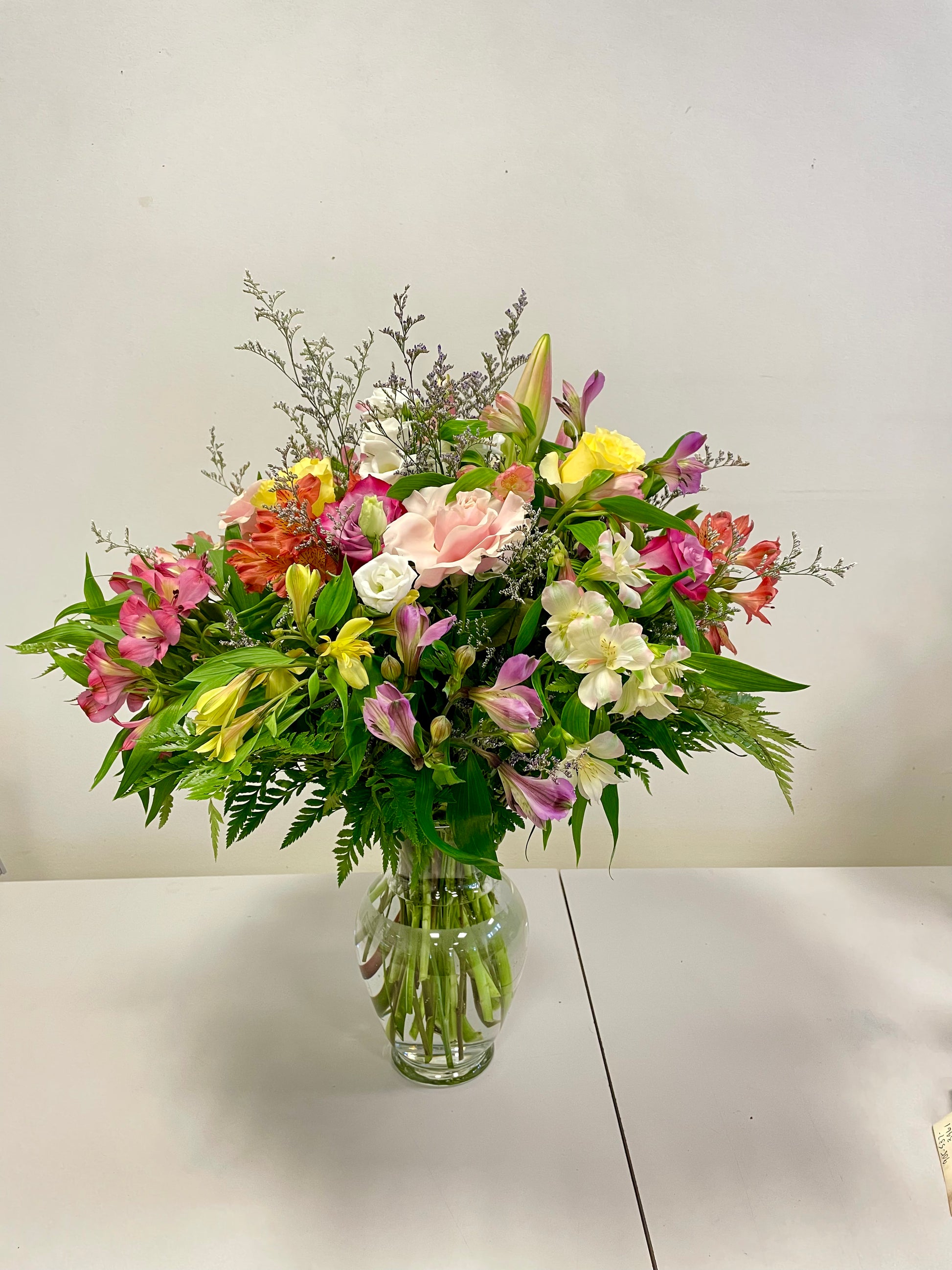 The Summer Serenade by Posies Flowers & Fashion features a clear glass vase filled with a vibrant bouquet of mixed flowers, including pink lilies, yellow and pink alstroemerias, and various green foliage. The arrangement is showcased against a plain white background on a white table. Please note that substitutions may be made based on flower availability.